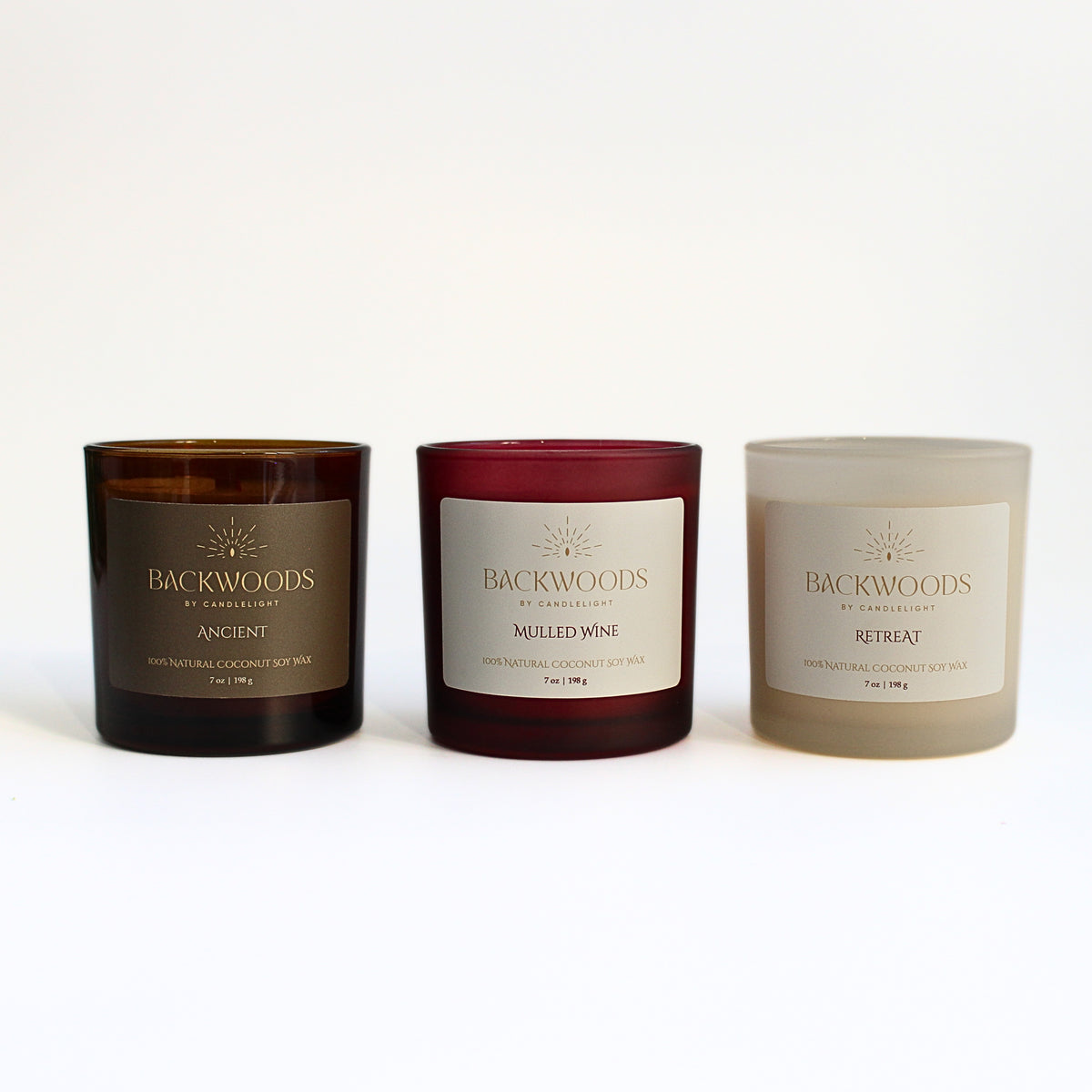 Backwoods By Candlelight fall scents: Ancient, Mulled Wine and Retreat 7 oz wooden wick candles side by side in amber, red and ivory glass jars