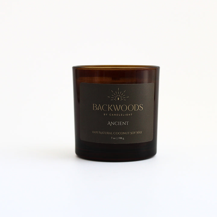 Ancient 7 oz wooden wick candle in an amber glass jar with an olive and gold label, on a white background