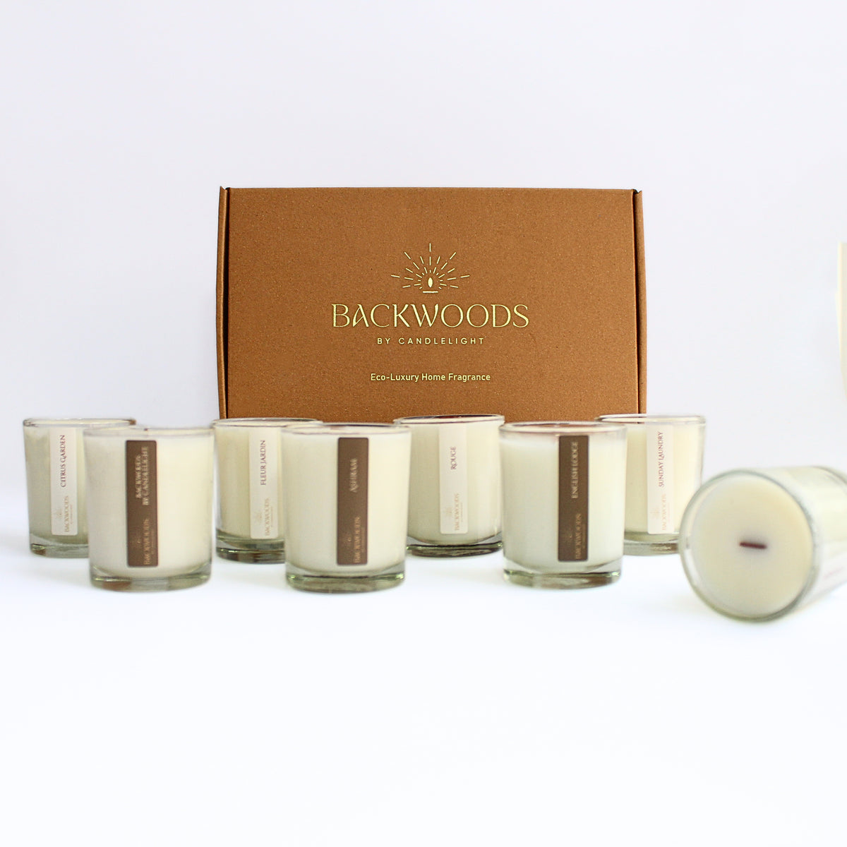 Complete Discovery Scent Set: eight 2.5 oz wooden wick votives lined up in front of a kraft Backwoods By Candlelight box with a gold foil logo