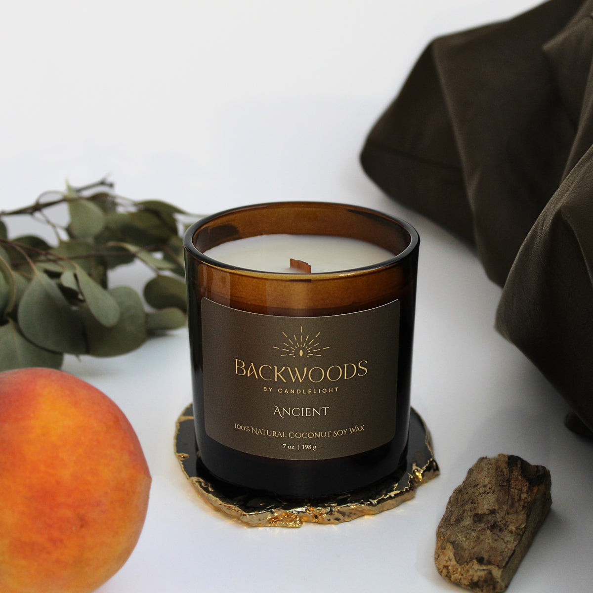 Ancient wooden wick candle in an amber glass jar on a gold-edged agate coaster, with a ripe peach, eucalyptus sprigs, sandalwood and dark linen