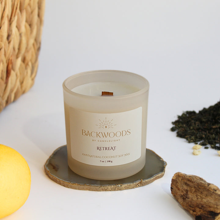 Retreat wooden wick candle in a frosted ivory jar on an agate coaster, with a fresh lemon, loose green tea and sandalwood beside a woven basket