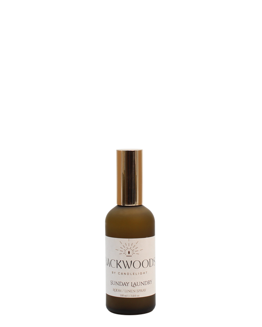 Brown spray bottle with gold cap labeled 'Backwoods by Candlelight Sunday Laundry' on a white background