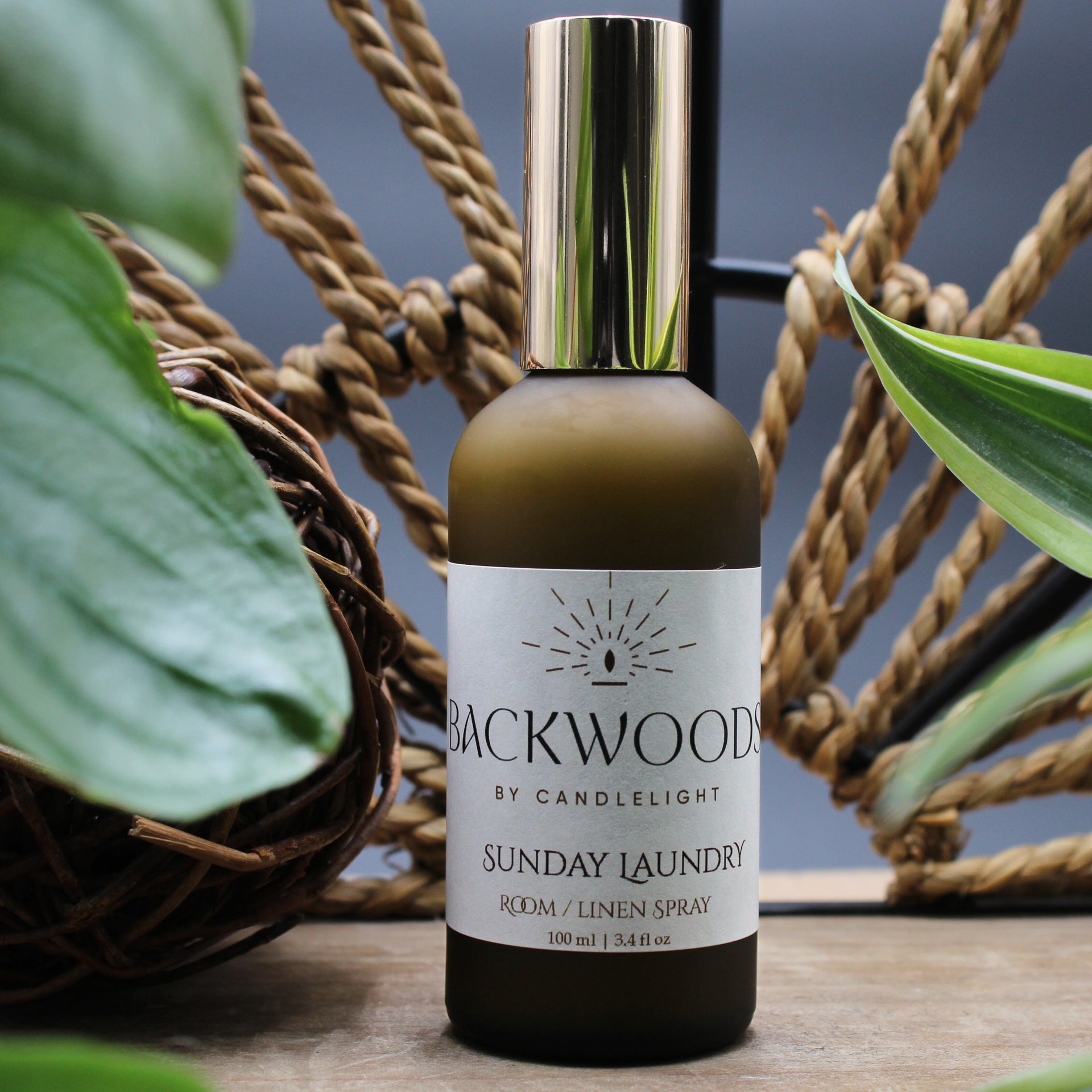 Brown spray bottle with gold cap labeled 'Backwoods by Candlelight Sunday Laundry' on a white 