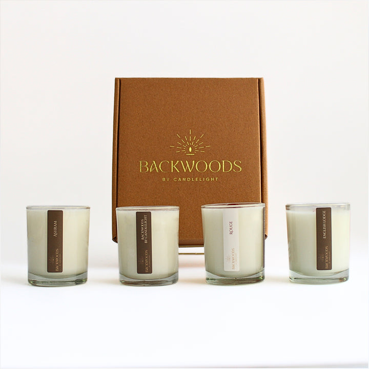 Warm & Cozy Discovery Scent Set: Ashram, Backwoods By Candlelight, Rouge and English Lodge 2.5 oz votives in front of a kraft box with a gold foil logo